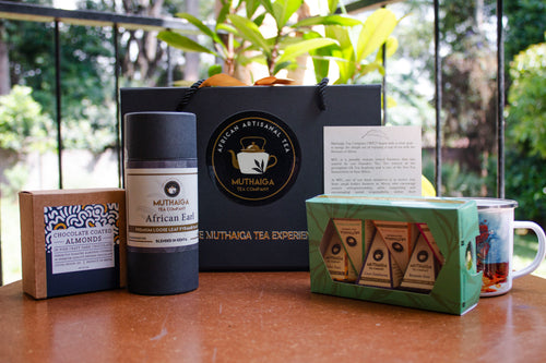 A Gift Hamper for Him in Kenya, including chocolates, tea bags, a tea cup, and a black logoed gift box, all displayed on a wooden surface with a plant in the background.