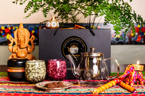 A Premium Diwali Gift Hamper Tea set with decorative items on a colorful tablecloth, featuring a black gift box with 'Dhama' branding, a teapot, ceramic cups and Swahli Oolong , Muthaiga Tea's Oolong blend tea.